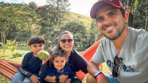 Foto da Família Pé de Pêra: Jonas, Rebeca e seus dois filhos, Miguel e Beatriz, sorrindo em um hotel fazenda de Camboriú sob o sol e sentados na rede.